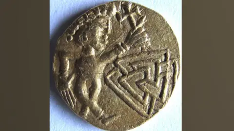 Adrian Marsden The gold coin. It depicts a male figure with an oversized head, with crossed legs and wearing a Roman-style headdress or diadem. Its right arm is down by its side and its left arm is raised up and appears to be holding a cross. Beneath the cross on the right is an interlocking design of three triangles. 