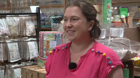 Catherine is wearing a pink top with brightly coloured balls stitched on her shoulder. She is standing in he farm shop with a range of packets of food behind her and a picture of ice cream behind her.