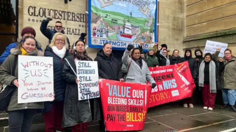 A group of striking NHS workers - perhaps around 20 people - stand in front of Gloucestershire's Shire Hall holding placards with slogans such as "Without us, your doctor would be guessing" and "Still striking, still standing, still strong".