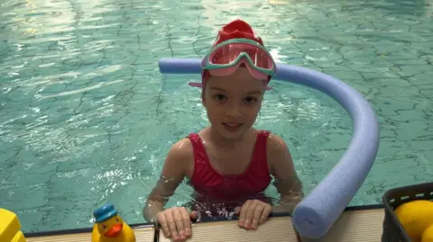 Maddie, a young girl, is in a red swimming suit, cap and goggles in a pool. There are pool noodles and a yellow duck near her.