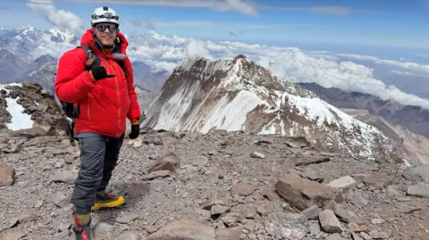 NDCS Michael Woods stands on the summit of a mountain with snowy peaks and clouds visible in the distance against a blue sky. He is wearing a white helmet, reflective sunglasses, a thick red coat and grey overtrousers. 