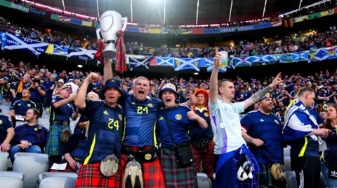Group of jubilant Scotland football fans in a packed stadium, many wearing kilts and navy shirts, cheering with arms raised; one holds a large inflatable trophy while others wave drinks and flags, with Scottish flags draped along the stands behind them.