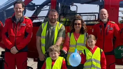 Lizzie Danton Lizzie, Alex and their children with the air ambulance team. The four of them are wearing neon yellow vests. They all stand in front of the air ambulance.