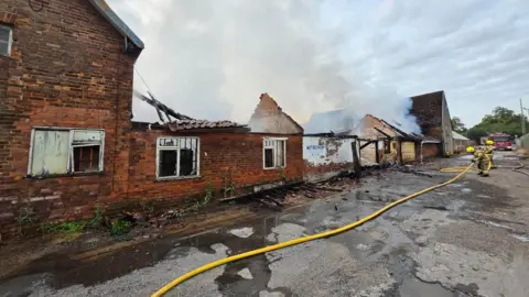 Nottinghamshire Fire and Rescue Service A damaged building with smoke blowing into the air. Firefighters are working in protective overalls.