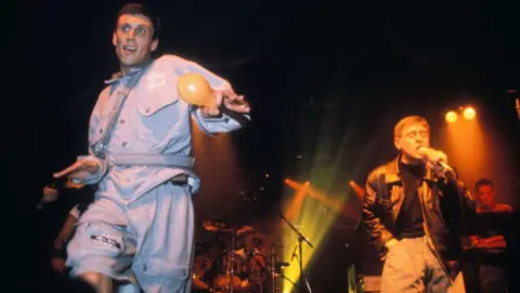 Getty Images Bez, in a light blue denim shirt and shorts, waves an orange maraca alongside Shaun Ryder, who wears a black T-shirt, black leather jacket and grey trousers while singing on an orange and green-lit stage with Happy Mondays