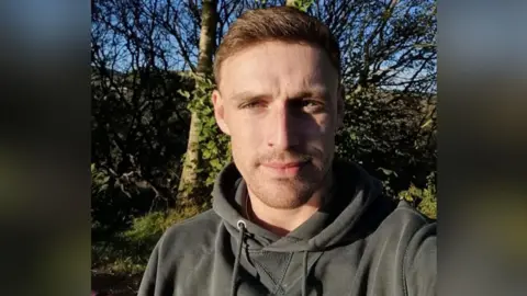 David Law looking straight at the camera. He is wearing a grey hoodie. He is stadning in front of a bare tree and some greenery. He has short, fair, brown hair and short stubble.