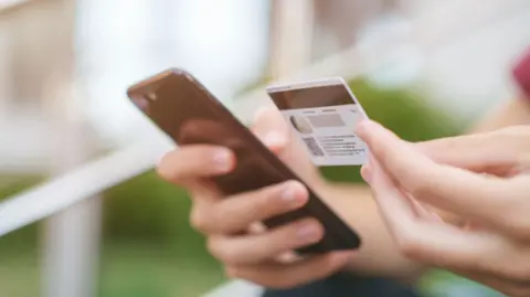 Getty Images An anonymous teenager holding a bank card in their left hand and a mobile phone in their right