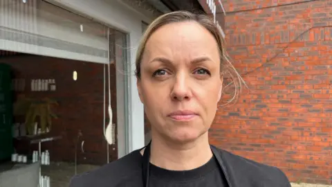 John Devine/BBC Jurga Ziliene is looking towards the camera and slightly frowning, wearing a black t-shirt. Behind her is a brick wall and an open garage door. 