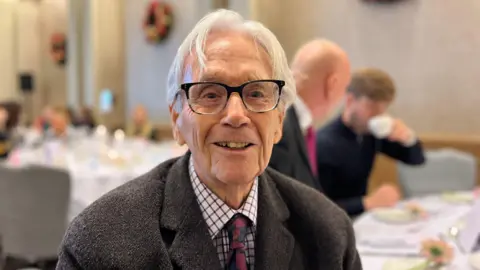 Terry Sadler smiling at the camera. He is wearing a suit and tie, glasses and has grey hair. In the background, you can see tables with formal place settings. 