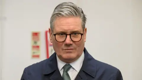 Reuters Picture of Keir Starmer - he has glasses on and a dark navy trench coat with a green tie.