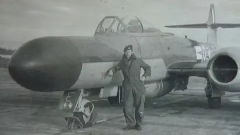 John Heys A black and white photo shows a young man in Royal Air Force overalls leaning on a jet plane which has a large, long nose.