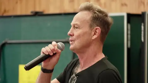 BBC Jason Donovan, wearing a black T-shirt, holding a microphone and singing into it.