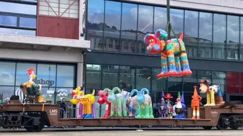 BBC Various large sculptures the Gromit sculpture with their own unique paintings. They are standing on a railway line, and one is being lifted up in the air by a crane outside the MShed in Bristol.