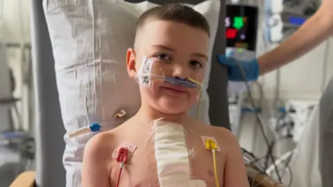 Máirtín McGabhann A young boy sitting in a hospital chair holding a football. He has a large bandage on his chest and several tubes and wires attached to his face and body.