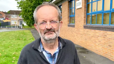 A man wearing round glasses with transparent frames in a dark grey jumper standing outside a brick building with grass and a tree in the background