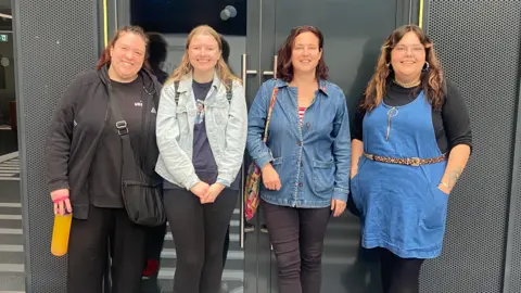 Gloucestershire Arts and Social Projects Four women standing outside a grey building. The lady on the left is wearing a grey hoodie and black trousers. The lady next to her on the right is wearing a denim jacket and black leggings. The lady on the right to her is wearing a denim jacket and black jeans and the lady on the very left is wearing a blue dress with a leopard print belt wrapped round. They are all smiling. 