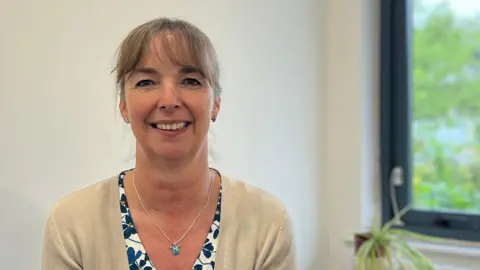 Mid shot of Lyn Gooding sitting in front of a pale wood desk which has a spider plant on it, a window and white wall. Lyn is smiling at the camera, has her brown hair tied back and is wearing a floral v neck top and tan coloured cardigan over it. She has a turquoise pendant necklace on.