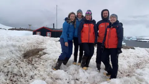 UK Antarctic Heritage Trust A picture of Dale Ellis wth her four other teams mates in Port Lockroy. They are dressed in outdoor weather clothing and are stood together smiling in the snow. 