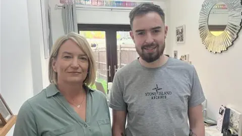 A woman in a green shirt and a man in a grey t-shirt, stood together in a hallway. Small pictures and a round mirror are hung on the walls.