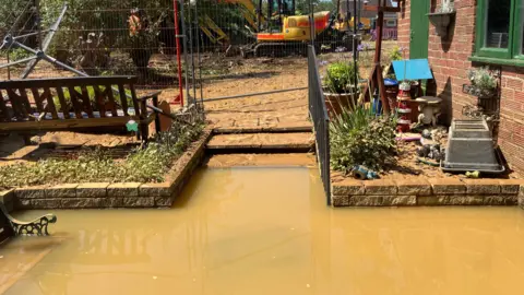 Kate Bradbrook/BBC A flooded garden, showing steps, a bench, a metal barrier, and plants. A large part of dirty water is in the garden.