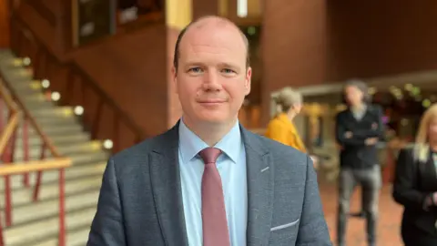 Gordon Lyons wearing a gray blazer, blue shirt and red tie