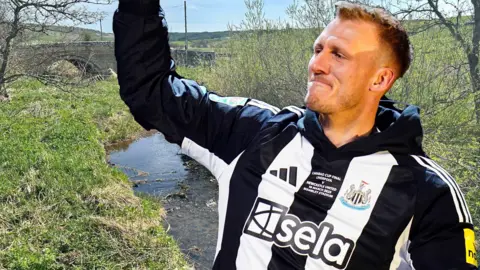 Footballer Dan Burn with his arm raised, and who is wearing the black and white Newcastle kit, is superimposed on to a photo of a small river on the Big Dan Burn Walk.