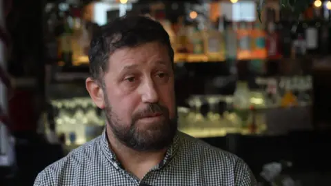 BBC / Peter Whittlesea A bearded man with dark hair and wearing a checked black and white shirt. He is standing inside a restaurant.