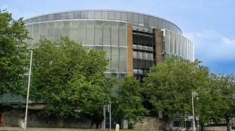 A circular shaped building with lots of windows. Trees are surrounding it.
