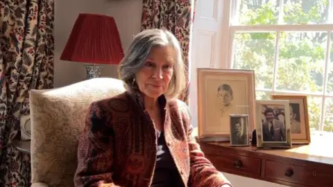 A woman with grey, white hair looks slightly off camera while sat in a light blue and white patterned chair. She has a red and black patterned jacket on over a dark top. To her left is a wooden chest of drawers with several family photos on top and a window behind that.