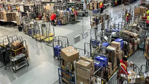 A busy sorting office  is filled with huge metal cages. There are boxes and parcels inside them.