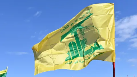 The yellow and green Hezbollah flag flies in the breeze, against a clear blue sky.