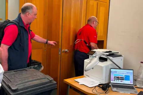 George Freeman Two men wearing red shirts leave an office after capturing a fox. The office has wooden doors and cupboards and a laptop can be seen on a desk