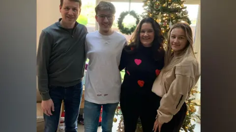 Yvonne Bruce Simon, Jack, Yvonne and Nadia Bruce stood in front of a Christmas tree. Simon is wearing a grey pullover and jeans, Jack is wearing a white t-shirt and jeans, Yvonne is wearing a black jumper with red hearts on it alongside black trousers, and Nadia is wearing black trousers and a beige hoodie.