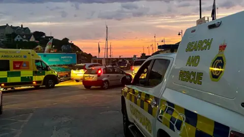 The image shows emergency vehicles, including a coastguard search and rescue truck and a South Western Ambulance Service ambulance, parked near Newquay Harbour at dawn. The sky is streaked with orange and pink light from the sunrise beneath dark clouds, while cars and harbour buildings can be seen in the background.