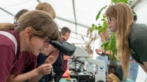 Royal Norfolk Show/James Bass Photography A teenager looks through a microscope, with other children in the background, and an instructor overlooking. Near them, inside a white marquee, is a plant.