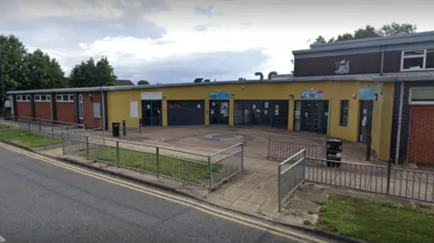 Google Holme Wood Library, in Bradford. Pictured is a yellow single-storey building behind a wide pavement.