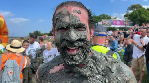 Richard Knights/BBC A laughing, bearded man with brown hair covered in mud with crowds behind him.