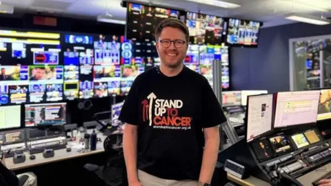 Cancer Research UK Jack wears a black Stand Up To Cancer shirt, and stands in the control room of a TV studio, backed by control desks and dozens of monitors. He has dark red hair and wears glasses.