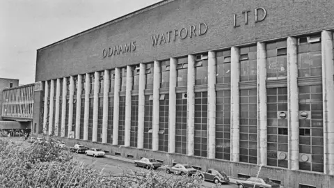 Getty Images A black and white photograph of the Odhams Watford printing plant.
The building's architecture is monumental and symmetrical, with a long facade dominated by tall, vertical columns and large multi-pane windows.