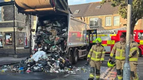 Refuse strewn on the road in the aftermath of a refuse wagon blaze with firefighters and a fire engine also in view