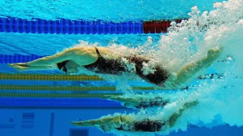 Generic image of two racing lane swimmers underwater. They both have arms outstretched in the water, wearing black swim suits and have underwater bubbles and froth extending behind them.