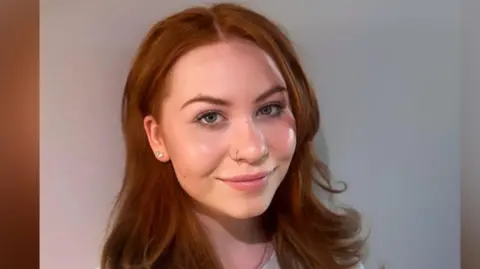 A professional headshot of Liliyana who is smiling to camera. She has long, slightly wavy brown to reddish hair and stud earrings. She is wearing a grey jumper