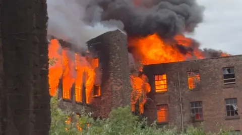 Image shows flames and smoke coming out of the red-brick, 19th-century Hotspur Press building in Manchester. 