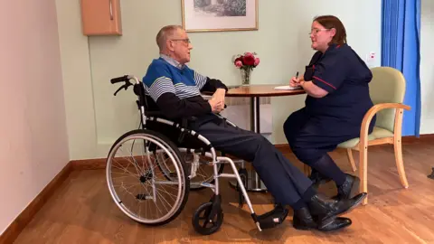 Man in a wheelchair and a nurse sitting at a table in discussion, while filling out a form. 