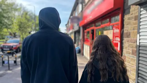 A boy in a black hoodie and a girl with curly brown hair look out a street, backs turned. To their right is a row of shops, to their left are trees and a car.