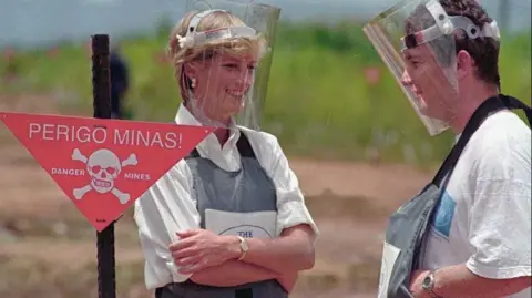 ANTONIO COTRIM/AFP via Getty Images Princess Diana wearing a heavy duty protection vest and face shield at a minefield in Angola, stood next to a man in the same outfit and behind a red sign that says danger mines with a skull and crossbones image.