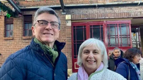 BBC David and Keren Leivesley smile as they pose together outside the Art Deco cinema entrance, which has red doors with lattice windows. He wears a dark coat and green scarf, while she wears a white coat.