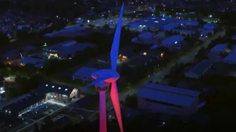 An aerial night time view of a large wind turbine illuminated with blue and pink lighting, rising above an industrial and residential area. Roads, buildings, parked vehicles, and scattered streetlights surround the turbine.