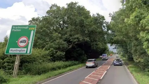 A rural road with cars travelling up and down on it. A roadsign to the side indicates Scarborough and Malton are straight ahead.