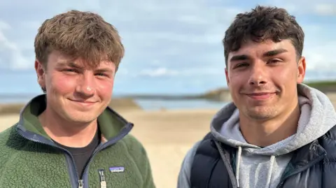 BBC Euan and Andrew stand side by side smiling at the camera, with a yellow beach and blue sea behind them. Euan has brown hair and is wearing a green fleece. Andrew has black hair and is wearing a grey hoody beneath a navy body warmer.
